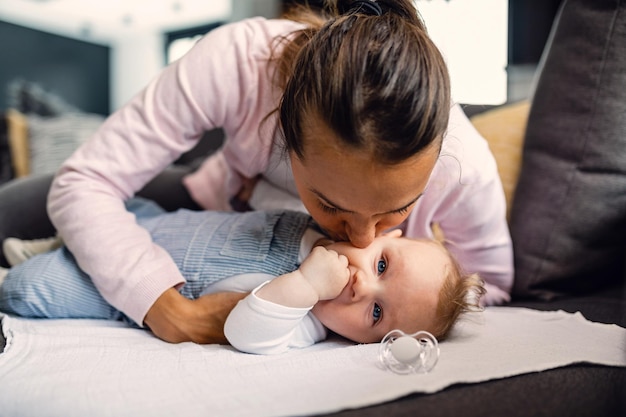 Young loving mother kissing her baby boy on cheek with affection while spending time with him at home