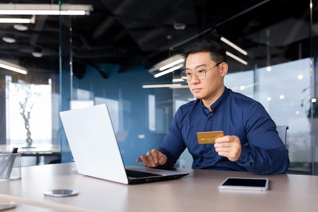 A young male asian banker works in the office at a laptop holds a credit problem in his hands