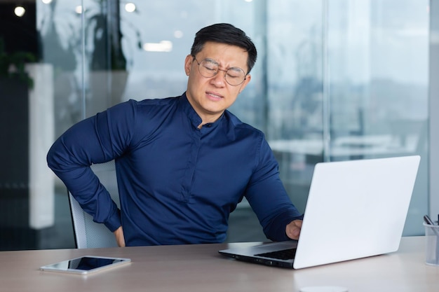 A young male asian student sits in the office at a desk with a laptop he feels severe pain in his