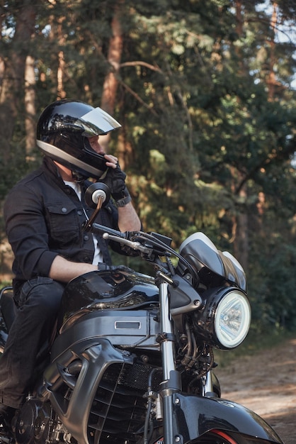 A young male biker travels on a motorcycle alone stopped and puts on a helmet on the side of a fores