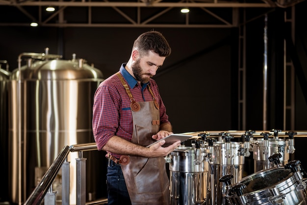 Photo young male brewer in leather apron supervising the process of beer fermentation at modern brewery fa...