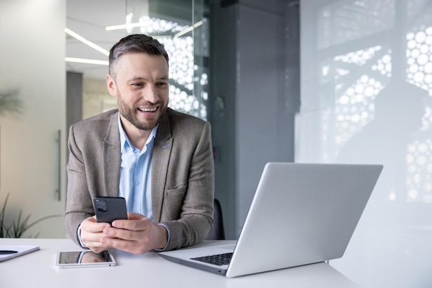 A young male businessman sits at a desk in the office holds a phone in his hands and communicates