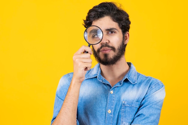 Young male detective holding magnifying glass by right eye while standing in front of camera in isolation