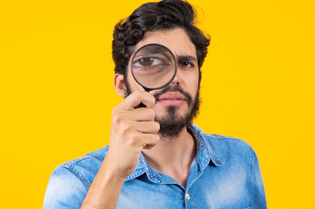 Young male detective holding magnifying glass by right eye while standing in front of camera in isolation