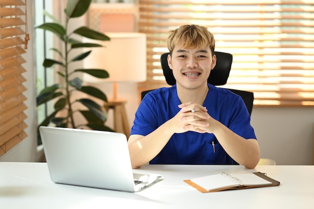 Young male doctor sitting front of laptop at his personal office and smiling to camera