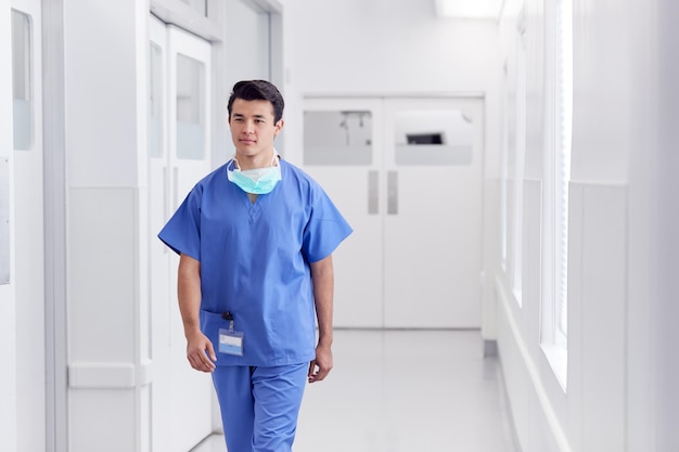 Young Male Doctor Wearing Scrubs Walking Along Hospital Corridor