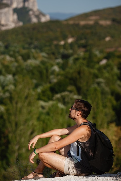 Young male hiker with backpack relaxing on top of a mountain during calm summer sunset