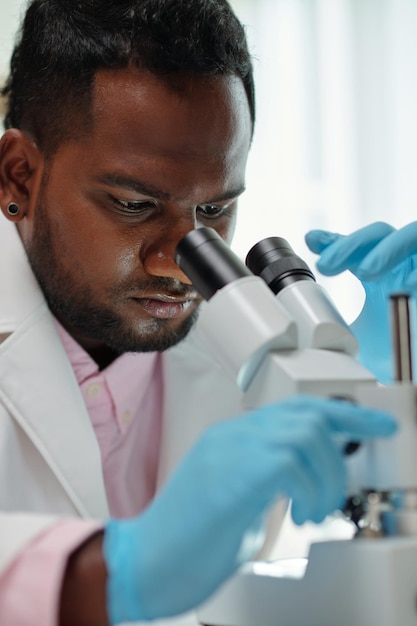 Young male microbiologist in protective gloves studying new virus or substance in microscope during