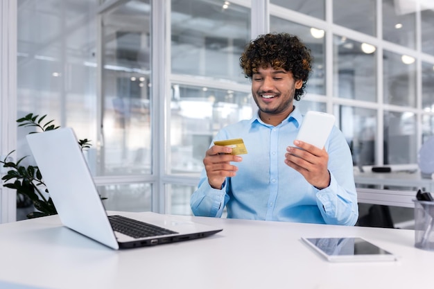 A young male muslim student sits in the office at a desk with a laptop and uses a credit card holds