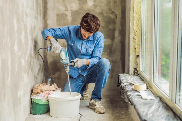 Young male painter kneads putty with water in a bucket using a hand-held mixer for building mixes