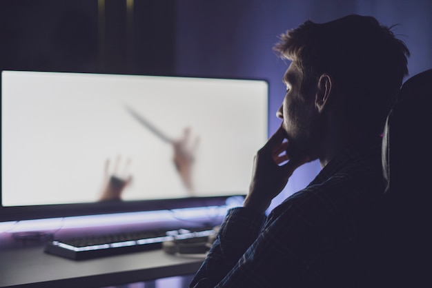 A young male person sitting in front of the screen and watching the horror movie at night, scary emotions