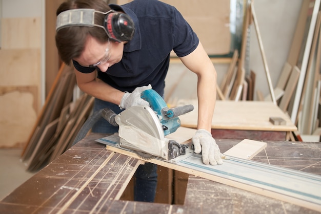 Young male in protective headphones and white gloves works on jigsaw in workshop