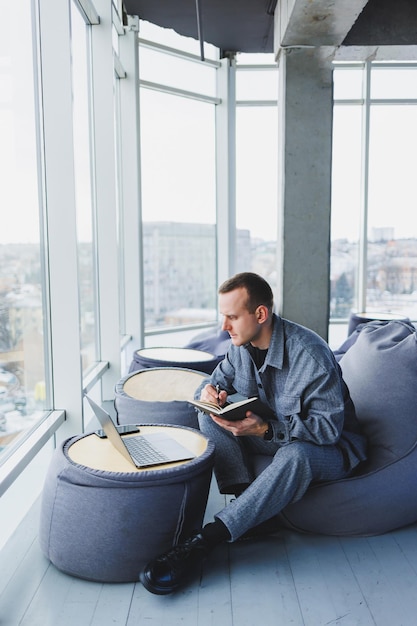 A young male student in a casual suit resting in a comfortable pouf while studying makes notes in a notebook an attractive freelancer in a suit works remotely