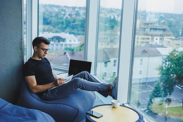 A young male student of Caucasian appearance sits in a bag chair and takes notes in a notebook The manager sits by the window and writes the work schedule