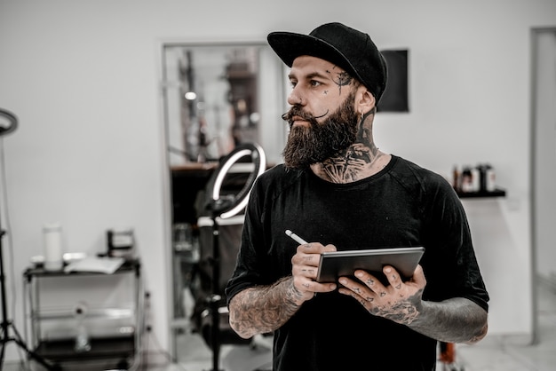 Young male tattoo artist with beard holding pencil and sketch looking positive and happy standing and smiling in workshop place.