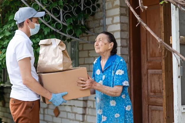 Young male volunteer in mask gives an elderly woman boxes with food near her house. The son helps a single elderly mother. Family support, caring. Quarantined, isolated. Coronavirus covid-19. Donation