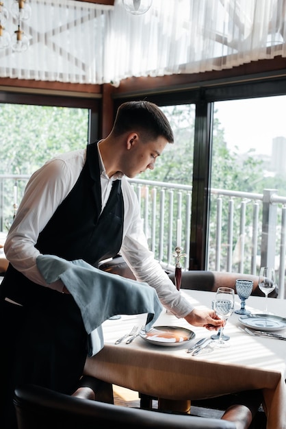 A young male waiter in a stylish uniform is engaged in serving the table in a beautiful gourmet restaurant. A high-level restaurant. Table service in the restaurant.