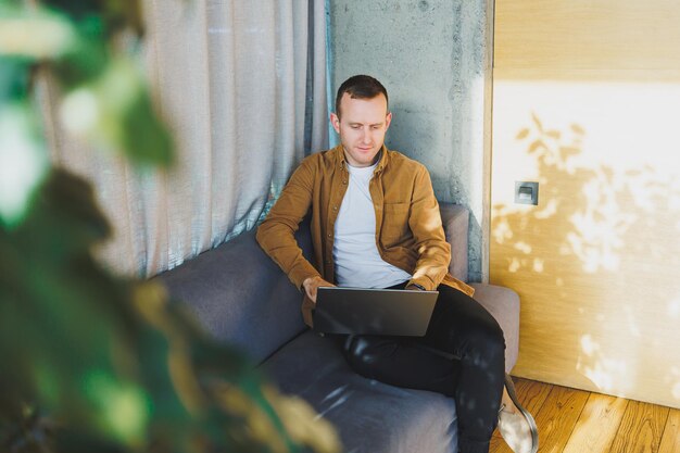 Young male worker in casual clothes and glasses looking at laptop while sitting on sofa with laptop in bright modern workspace A successful freelancer works remotely online in a workspace