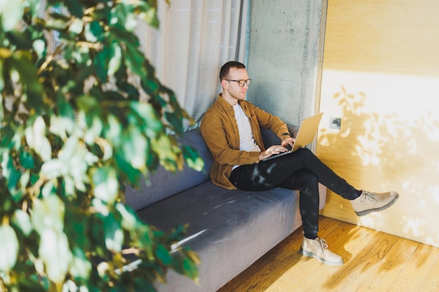 Young male worker in casual clothes and glasses looking at laptop while sitting on sofa with laptop in bright modern workspace A successful freelancer works remotely online in a workspace