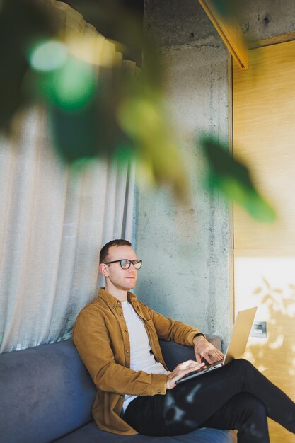 Young male worker in casual clothes and glasses looking at laptop while sitting on sofa with laptop in bright modern workspace A successful freelancer works remotely online in a workspace