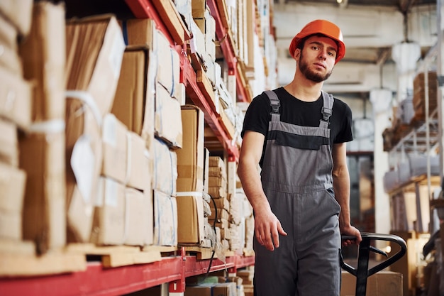 Young male worker in uniform is in the warehouse pushing pallet truck.