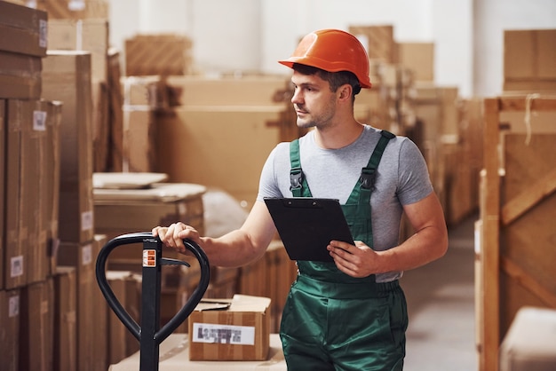 Young male worker in uniform is in the warehouse with notepad and pallet truck.