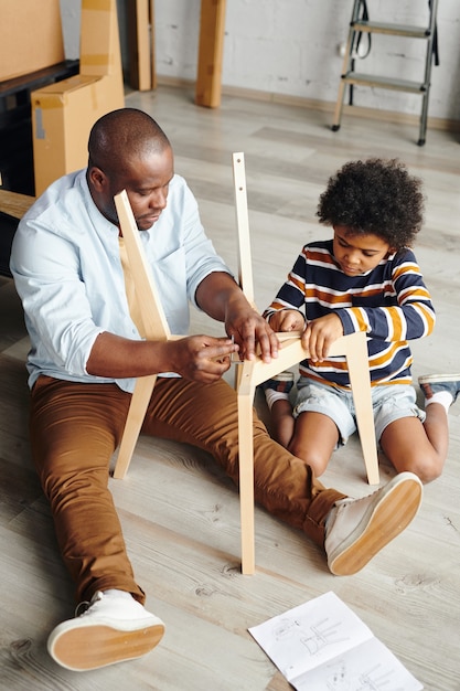 Young man of African ethnicity teaching his cute little son how to assemble new furniture while sitting on the floor of new flat or house