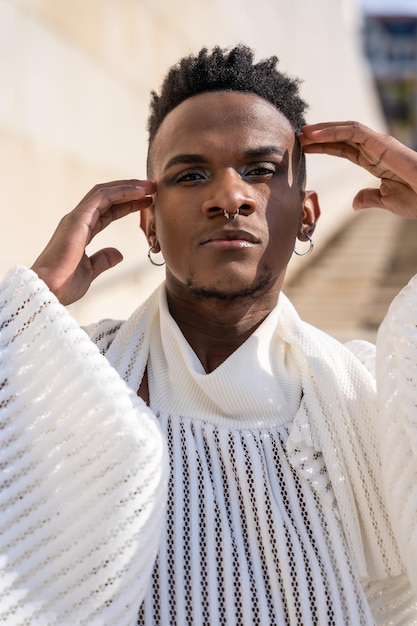 A young man of African ethnicity in a white contemporary suit and black leather boots stylish model on the street in the city on a brick wall background portrait with hands on his face seductive look