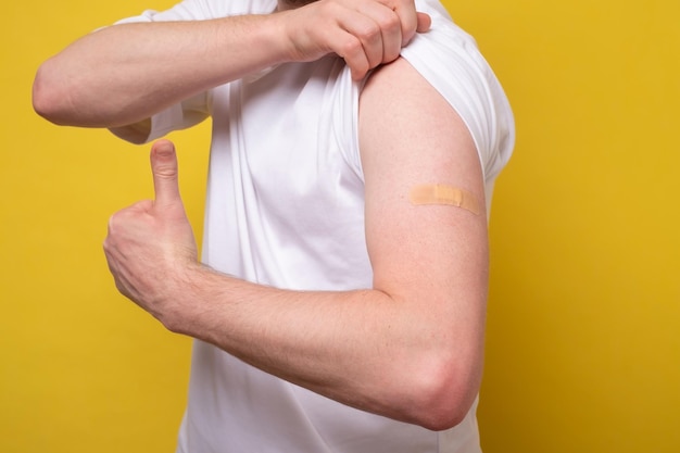 Young man after getting receive anti virus vaccine covid19 Showing arm and thumb up gesture Studio shot on yellow wall