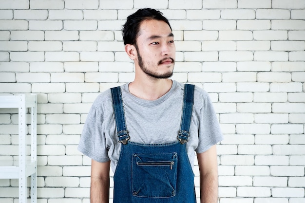 Young man in apron standing and smiling looking to outside in front of white brick wall.