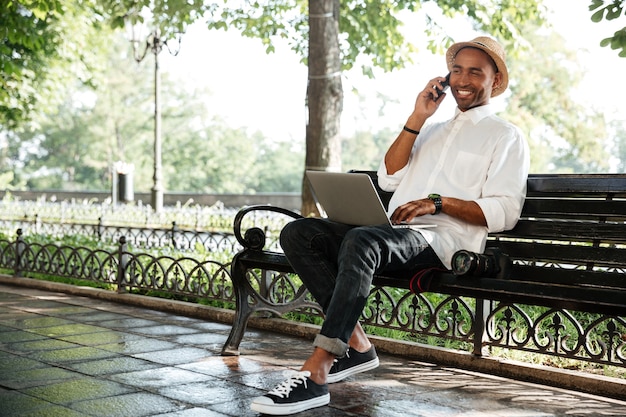 Young man on a bench with laptop and camera, talking with smartphone