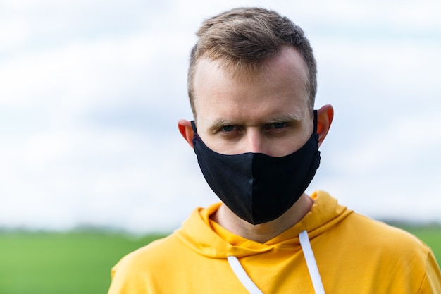 Young man in black protective antiviral mask on the street. The guy is resting in the park on a sunny summer day in a mask made by his own hands. Virus protection. Quarantine measures