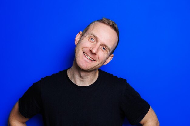 A young man in a black Tshirt on a blue background shows different emotions on his face Cheerful man with a smile Place for text Male beauty