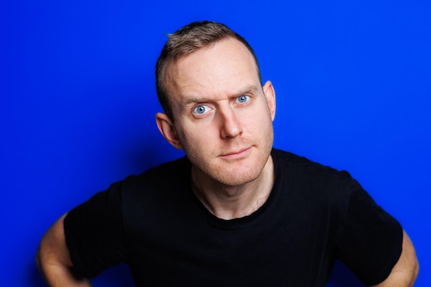 A young man in a black Tshirt on a blue background shows different emotions on his face Cheerful man with a smile Place for text Male beauty