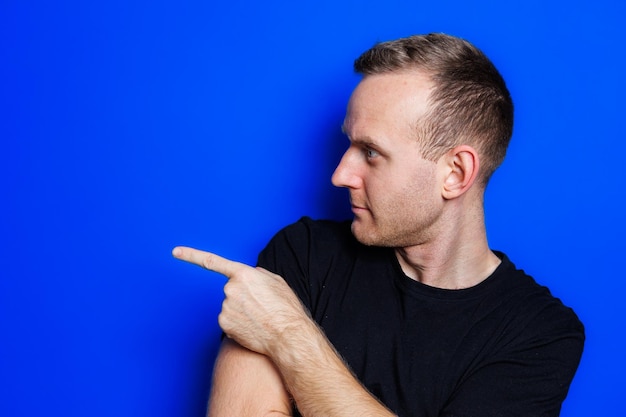 A young man in a black Tshirt on a blue background shows different emotions on his face Cheerful man with a smile Place for text Male beauty