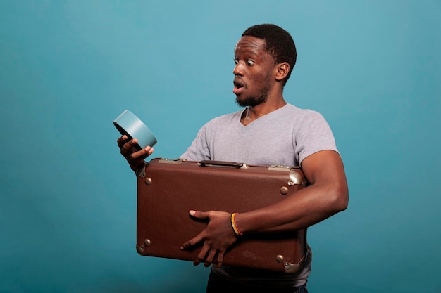 Young man carrying suitcase and running late for vacation trip, looking at time on clock over blue background. Person with retro briefcase having watch to prepare for work voyage.