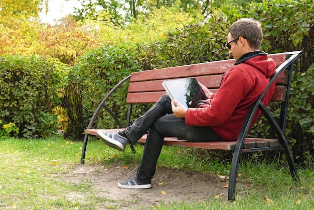 A young man in casual clothes in a warm jacket working outdoors in the autumn Park read a book on ultrabook teenager searching media content together on line with laptop sitting on bench in a park