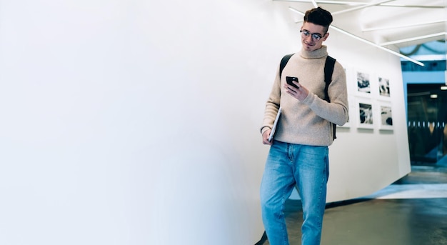 Young man in casual outfit and glasses holding laptop and using smartphone while standing