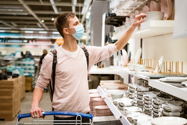 A young man of caucasian ethnicity in a protective mask and in casual clothes chooses the dishes in ...