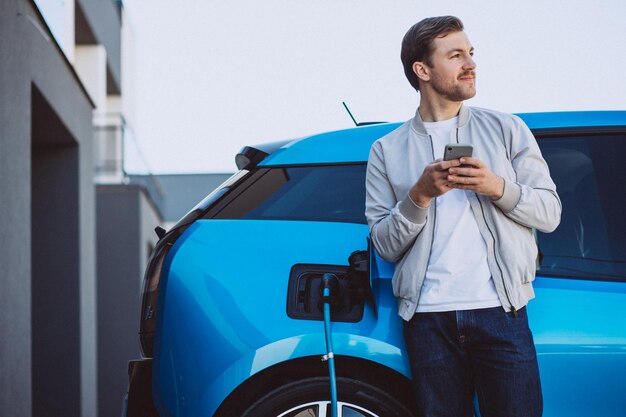 Young man charging electric car at home and using mobile phone