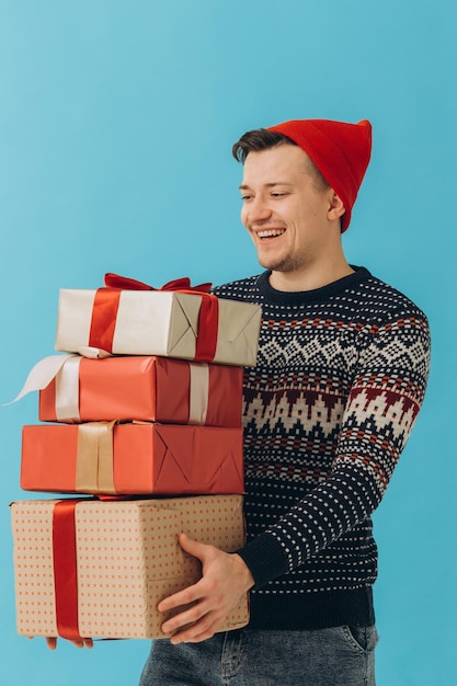 Young man in Christmas sweater and red hat hold many present boxes with gift ribbon bow isolated on blue background Happy New Year celebration conceptxA