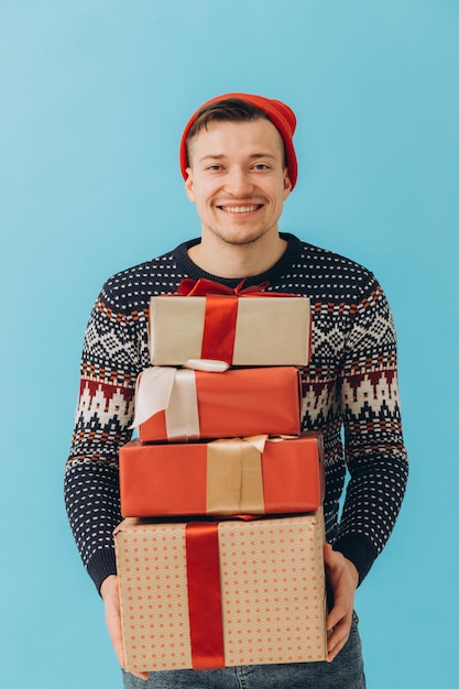 Young man in Christmas sweater and red hat hold many present boxes with gift ribbon bow isolated on blue background Happy New Year celebration conceptxA