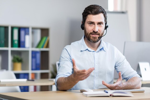 A young man a consultant an office worker a manager talking on a video call in headphones waving his arms explaining something Sitting at a computer desk in the office in a white shirt