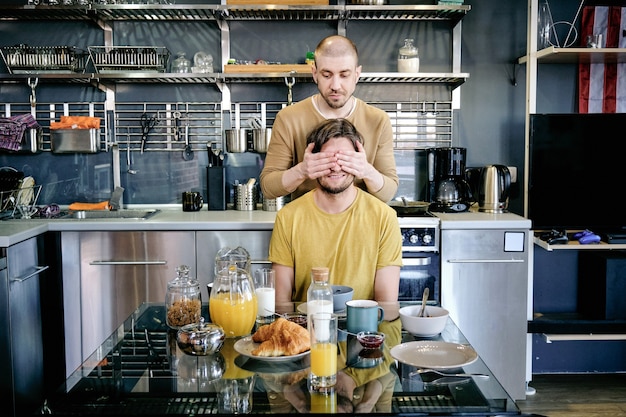 Young man covering eyes of his husbnd by hands by kitchen table