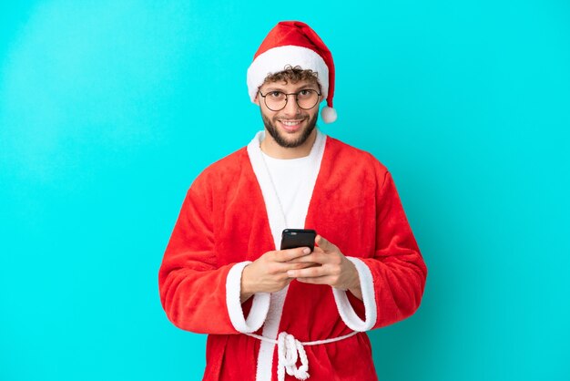 Young man disguised as Santa Claus isolated on blue background sending a message with the mobile