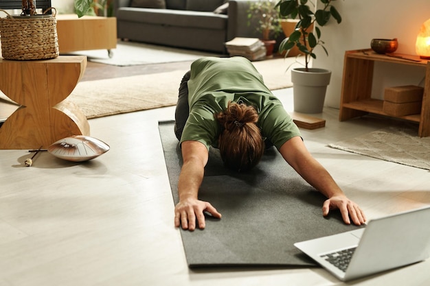 Young man doing stretching exercises on exercise mat while watching lesson online on laptop