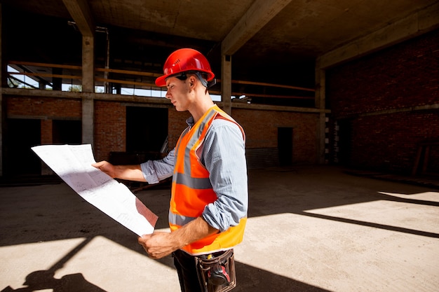 Young man dressed in shirt, orange work vest and helmet explore construction documentation on the building site inside the building under construction .