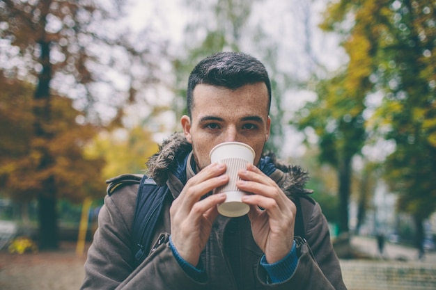 Young man drink takeaway coffee or tea outside in park or on street. Look straight on camera and pose. Autumn time. Cold outside