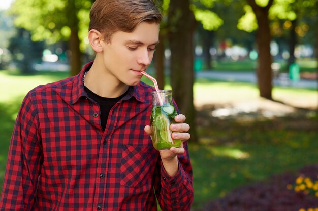 Young man drinks healthy detox tea with juice on green park background.