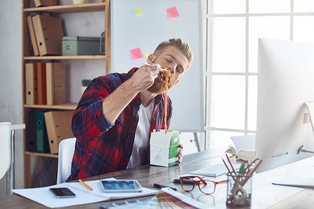 Young man enjoying asian food from delivery while sitting at his workplace in the office. Lifestyle concept